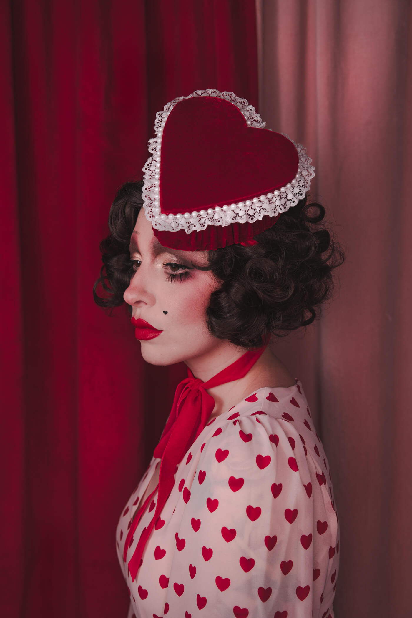 Pale girl with dark 20s 30s style hair and makeup, wearing a red velvet heart shaped box hat